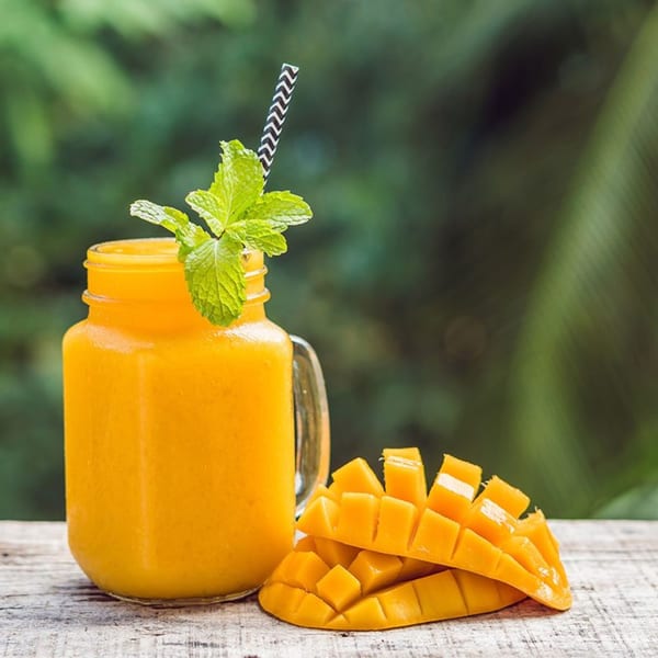 A Fudenjuce mango smoothie in a jar with mint and a striped straw, next to sliced mango.