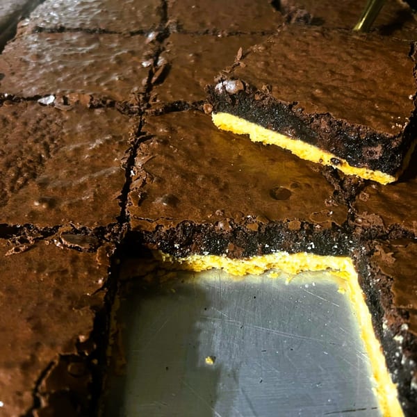A pan of Fudenjuce brownies with a golden crust, one piece removed revealing the bottom layer.